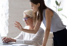 Two women, one seated at a desk with a laptop, the other leaning over her shoulder with her arm reached out as they speak, looking at the same laptop.