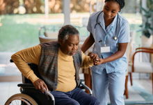 Young nurse assists a man to get up from wheelchair at a clinic. 