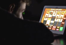 A man seen from behind over his shoulder, sitting in the dark, watching a digital slot game on a tablet.
