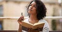 A woman with dark, curly hair holding an open, brown notebook in one hand, a pen held raised in the other.