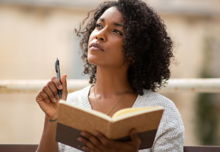 A woman with dark, curly hair holding an open, brown notebook in one hand, a pen held raised in the other.