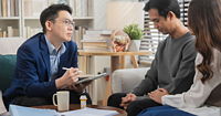A therapist holding a clipboard sitting in front of a couple, one of which is holding the othe's arm.
