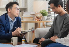 A therapist holding a clipboard sitting in front of a couple, one of which is holding the othe's arm.