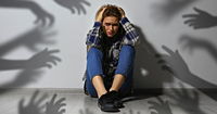 A scared woman sits near a wall. Shadows of hands reaching for her symbolize anxiety 