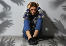 A scared woman sits near a wall. Shadows of hands reaching for her symbolize anxiety 