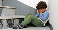 Little child with brown hair hiding in a corner with their face in their hands.