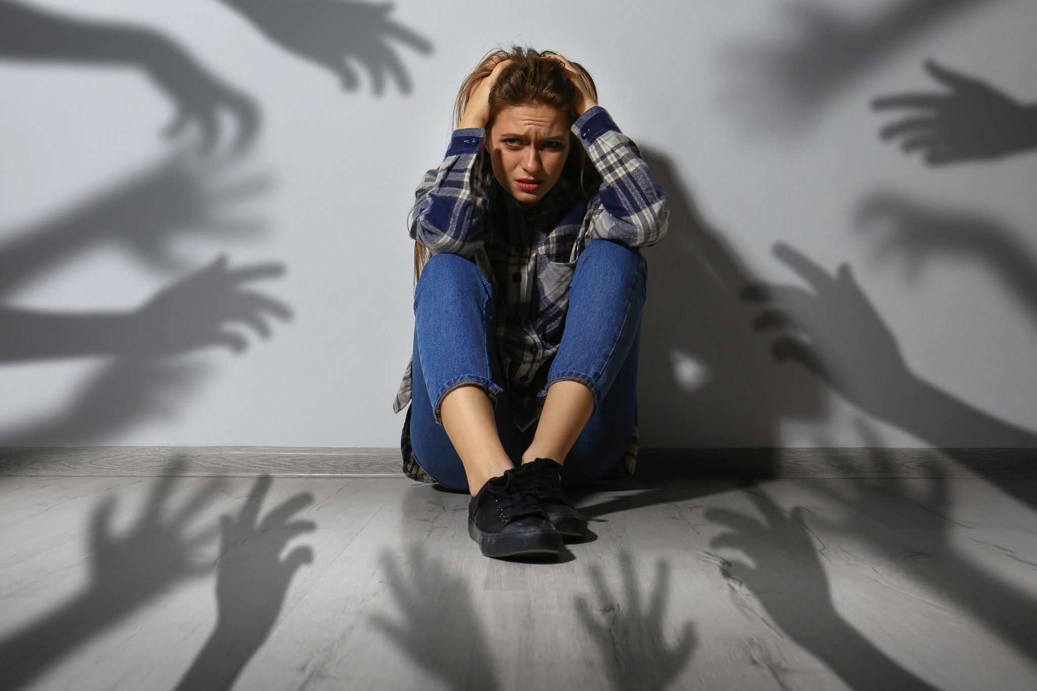 A scared woman sits near a wall. Shadows of hands reaching for her symbolize anxiety