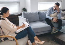 A therapist sitting in a chair at an angle from her client who has his forehead against one hand.