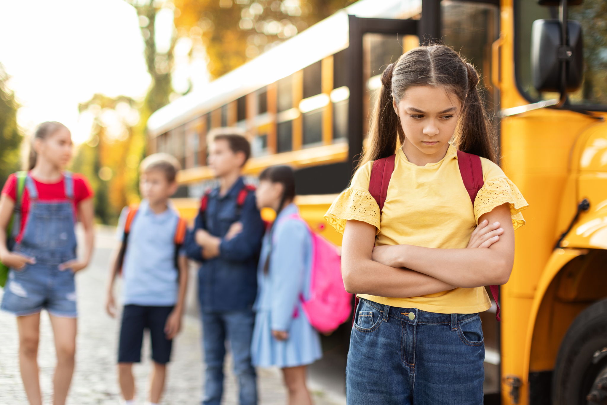 Singled out girl standing by herself next to yellow school bus.