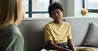 Young child sitting on couch interacting with therapist holding clipboard engaging in conversation inside a cozy living room during therapy session 