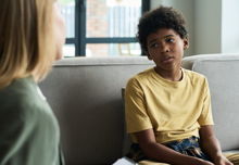 Young child sitting on couch interacting with therapist holding clipboard engaging in conversation inside a cozy living room during therapy session 