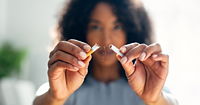 A woman with dark skin holding up a cigarette which has been broken in half. Her face is blurred in the background, putting focus on her hands.