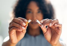 A woman with dark skin holding up a cigarette which has been broken in half. Her face is blurred in the background, putting focus on her hands.
