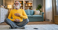 An older man in a yellow sweater and heaphones sitting cross legged on a white carpet, his hands pressed to his stomach and chest.