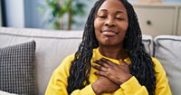 A black woman with long braids with her hands over her chest wearing a yellow sweater.
