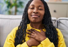 A black woman with long braids with her hands over her chest wearing a yellow sweater.