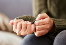 A pair of hands clenched together resting on the person's knees. The person is wearing a green sweater and worrying at the cuff.