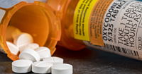 A bottle of oxycodone with a blurred hand holding some white pills in the background.