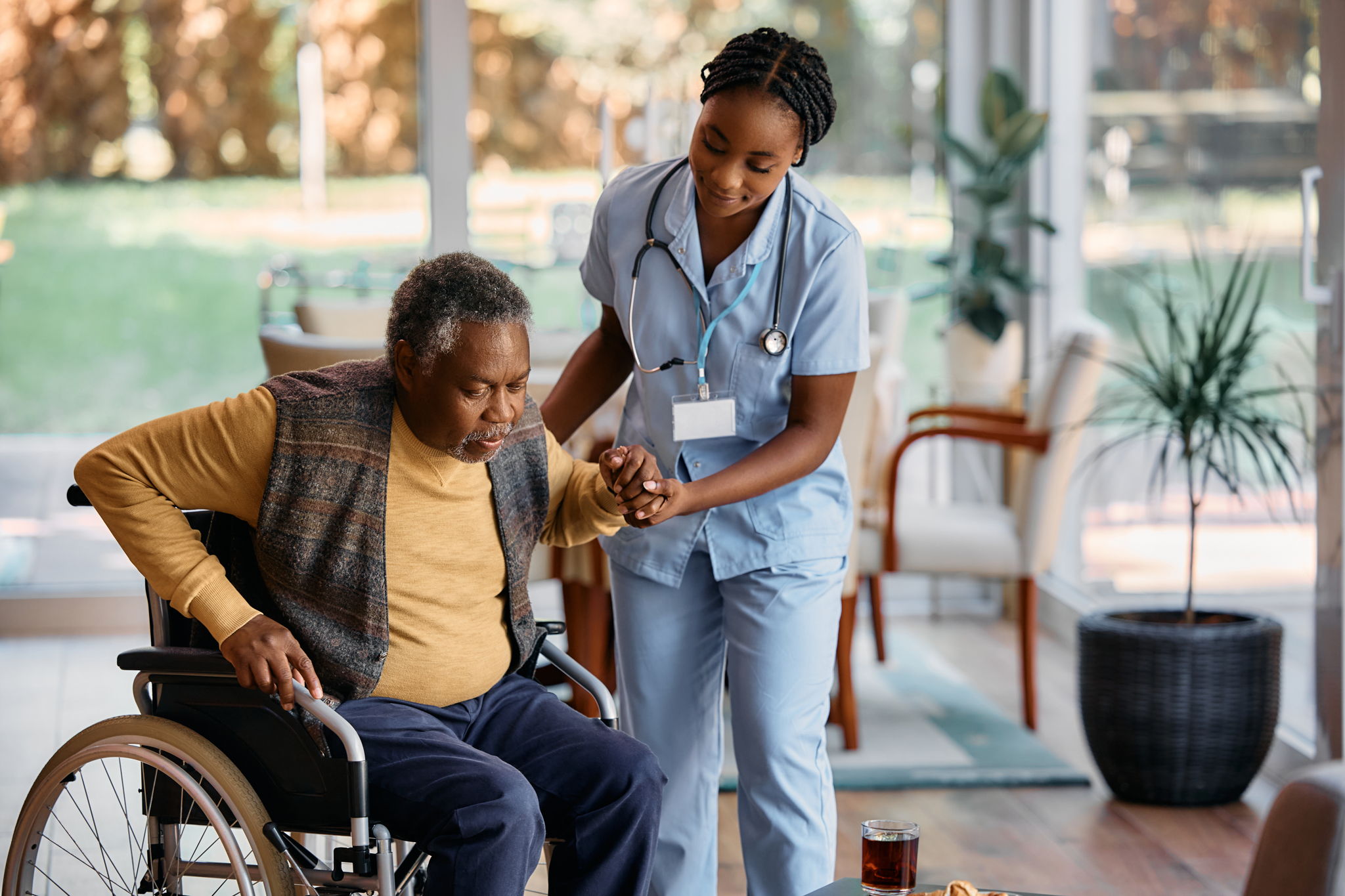 Young nurse assists a man to get up from wheelchair at a clinic.