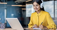 A smiling woman in a yellow shirt wearing white headphones, taking notes at her laptop.