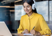 A smiling woman in a yellow shirt wearing white headphones, taking notes at her laptop.