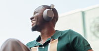 A black man pictured from below, looking up and smiling. He is wearing grey headphones and wearing a green polo short sleeve shirt with tan stripes on the chest.