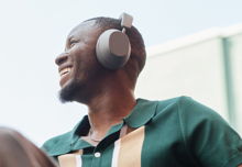 A black man pictured from below, looking up and smiling. He is wearing grey headphones and wearing a green polo short sleeve shirt with tan stripes on the chest.