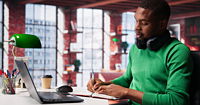 A younger black man wearing a green sweater with headphones around his neck taking notes at his laptop.