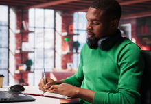 A younger black man wearing a green sweater with headphones around his neck taking notes at his laptop.