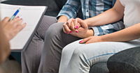 A view of a couple from the chest down, sitting on a couch, holding hands.