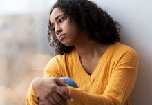 A woman with dark curly hair and a yellow sweater leaning against a window lookinf forlorn.