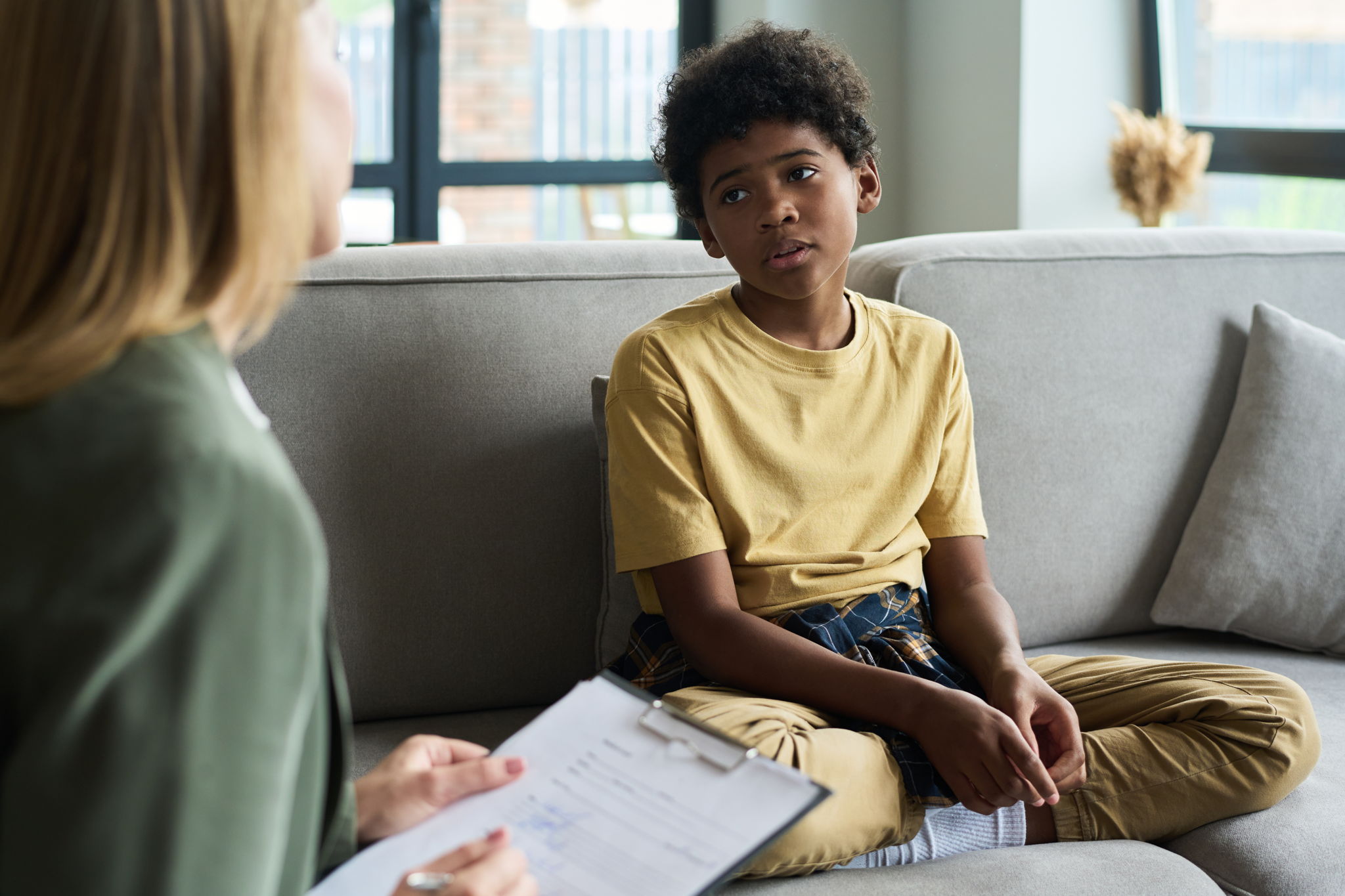 Young child sitting on couch interacting with therapist holding clipboard engaging in conversation inside a cozy living room during therapy session