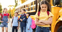 Singled out girl standing by herself next to yellow school bus.