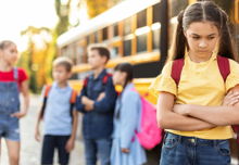 Singled out girl standing by herself next to yellow school bus.