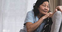 An older asian woman leaning against the back of a couch with her head resting on one arm.