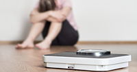 A bathroom scale in foreground, in background a woman hunched over and sad